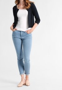 Cárdigan azul marino oscuro sobre una camiseta sin mangas blanca, jeans ajustados de mezclilla azul claro y zapatillas de pala rosa pálido. Tejido suave y diseño ajustado.