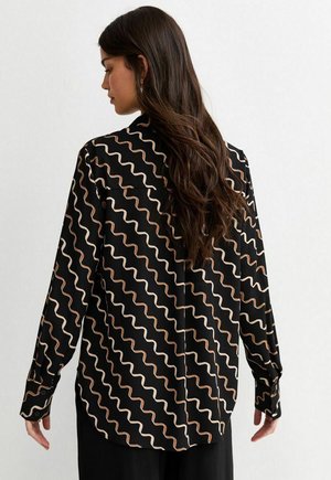 Vrouw met lang bruin haar, gekleed in een zwarte blouse met witte en bruine golvende strepen, gezien van achteren tegen een effen witte achtergrond.