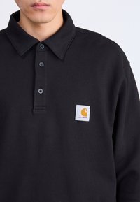 Polo shirt nera in cotone, con colletto classico, tre bottoni scuri e un'etichetta bianca con un logo arancione sul lato sinistro.