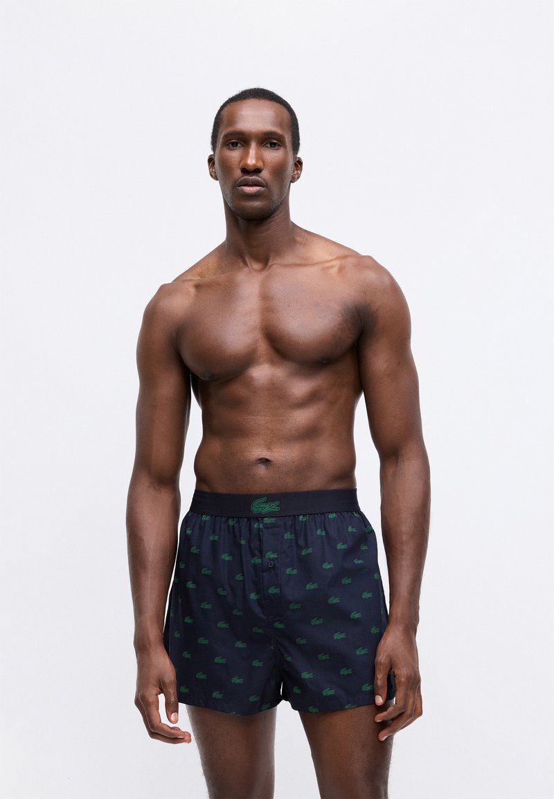 Navy boxer shorts featuring a green lacoste logo pattern, elasticated waistband, and a relaxed fit. Made from breathable fabric.