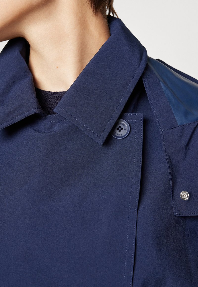 Trench-coat bleu marine avec un large col, fermeture par boutons et texture lisse. Présente des accentuations brillantes aux épaules et un détail de bouton logo.