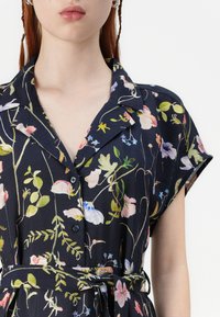 Blomstrete skjortekjole i marineblått, med korte ermer, krage, knappelukking og belte i midjen med blomster i forskjellige farger.