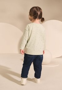 Svetlozelená košeľa s dlhým rukávom a malými vzormi, skombinovaná s tmavými denimovými nohavicami a bielymi topánkami. Dieťa vidieť zozadu, stojace na neutrálnom pozadí.