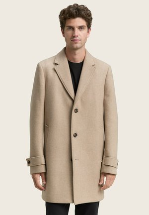 Hombre con abrigo beige abotonado sobre camisa negra, de pie frente a un fondo claro y sencillo, mirando hacia adelante con expresión neutral.