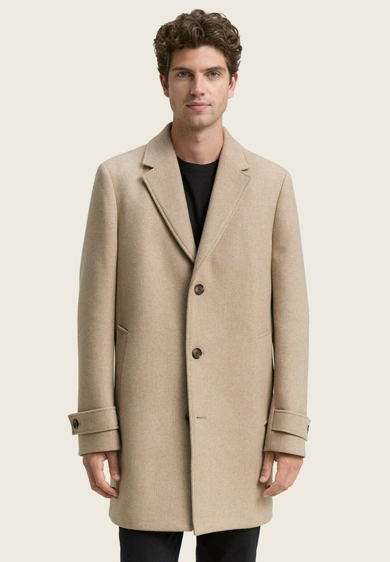 Hombre con abrigo beige abotonado sobre camisa negra, de pie frente a un fondo claro y sencillo, mirando hacia adelante con expresión neutral.