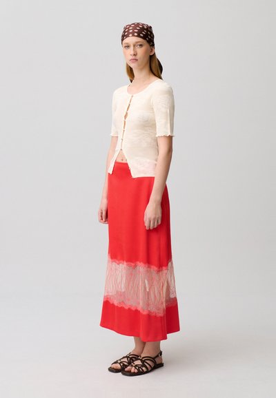 Donna che indossa una maglia crema a maniche corte con bottoni, gonna lunga rossa con pannello in pizzo, foulard marrone con motivi e sandali neri con cinturini, ferma in piedi.