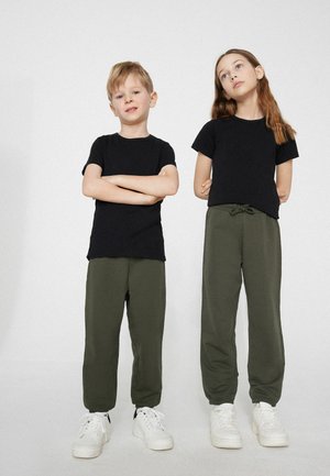 Deux enfants, un garçon et une fille, se tiennent côte à côte portant des t-shirts noirs, des pantalons vert olive et des baskets blanches sur un fond uni.