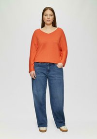 Oranje gebreide trui met een V-hals, gecombineerd met losse blauwe denim jeans. Model draagt lichte schoenen en staat tegen een effen achtergrond.