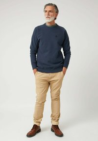 Sudadera navy con cuello redondo, puños acanalados y un pequeño emblema. Pantalones chinos beige ajustados, combinados con botas marrones de cordones.