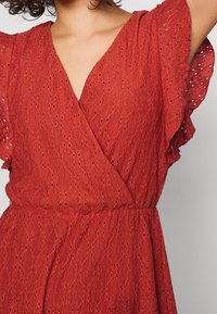 Robe rouge avec un design à enveloppe et des manches flottantes. La texture est légère avec des motifs œillets et une taille ajustée.