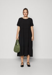 Crna haljina na više slojeva s kratkim rukavima, izrađena od mekog materijala. Model drži teksturiranu zelenu torbu i nosi crne balerinke. Jednostavan dizajn.