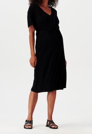 Black maternity dress with a V-neck and short sleeves, gathered under the bust. Made of soft fabric, knee-length, paired with black sandals.