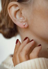 Złote kolczyki z kwiatowym wzorem, z trzema kryształowymi akcentami, łańcuszkowym zawieszeniem i małym zwisającym charms'em. Widoczny ton skóry w pobliżu.