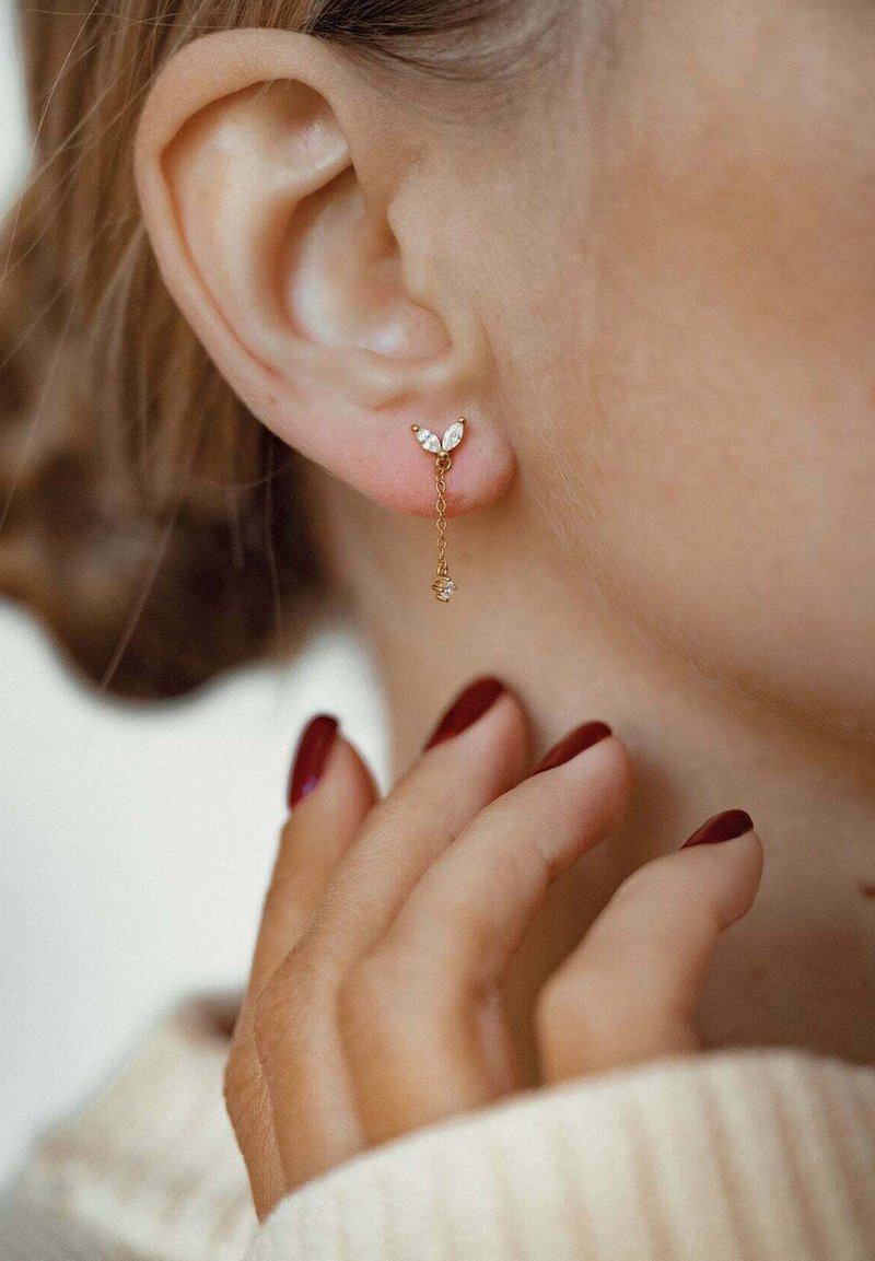 Złote kolczyki z kwiatowym wzorem, z trzema kryształowymi akcentami, łańcuszkowym zawieszeniem i małym zwisającym charms'em. Widoczny ton skóry w pobliżu.