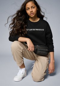 Sudadera negra con el logo blanco "Emporio Armani", pantalones jogger beiges, zapatillas blancas y un pouch negro con cremallera. Cabello rizado castaño.