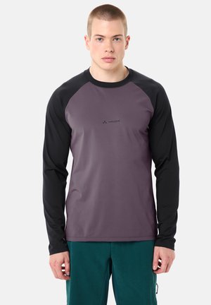 Giovane uomo con capelli corti che indossa una maglietta a maniche lunghe viola e nera e pantaloni verde acqua, in piedi e rivolto verso l'obiettivo su uno sfondo neutro.