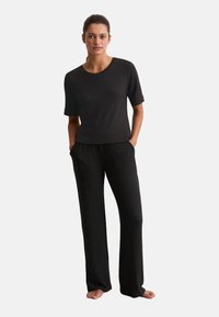 Mujer de pie, descalza, vistiendo una camiseta negra de manga corta y pantalones negros sueltos, con las manos en los bolsillos, frente a un fondo blanco.