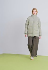 Giacca imbottita in verde chiaro, con colletto alto e texture trapuntata, abbinata a pantaloni cargo larghi verde scuro e sneakers beige.