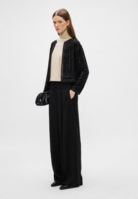 Μαύρο cropped μπουφάν με κατακόρυφες διακοσμήσεις, συνδυασμένο με απαλό καφέ ζιβάγκο. Χαλαρές μαύρες παντελόνες και μια μαύρη τσάντα συμπληρώνουν την εμφάνιση.