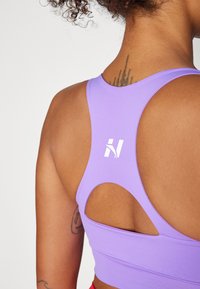 Top corto atlético morado con diseño de espalda cruzada, un corte abierto y un logo blanco en la parte trasera, fabricado con un material suave y elástico.