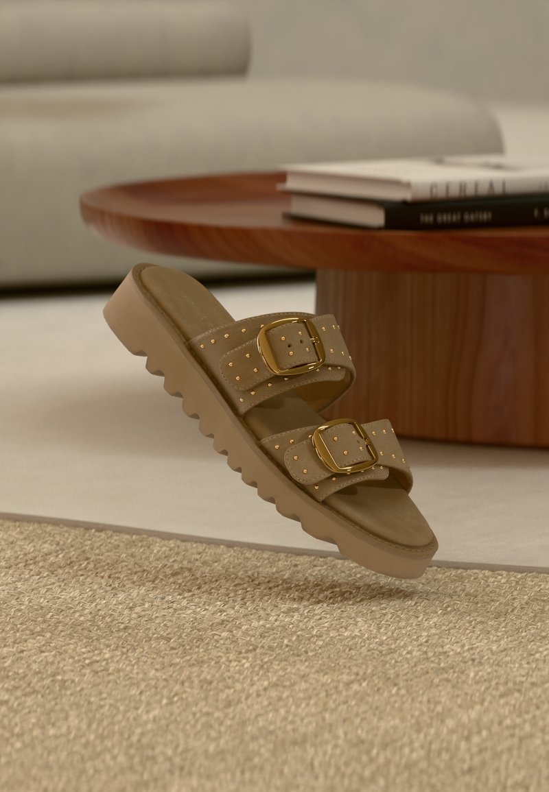 Sandale à glissière marron avec deux boucles dorées et détails cloutés, posée près d'une table en bois avec des livres empilés et un canapé beige en arrière-plan.