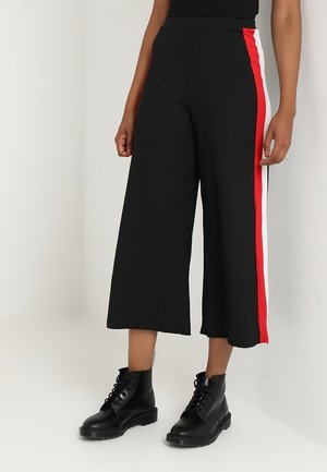 Persoon draagt zwarte wijde cropped broek met rode en witte zijstrepen en zwarte veteranklelaarzen.
