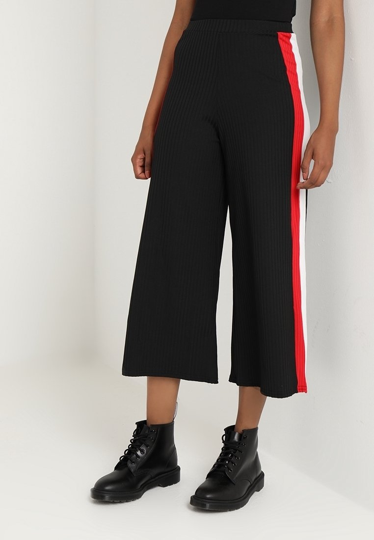 Personne portant un pantalon noir large et court avec des bandes latérales rouges et blanches, ainsi que des bottines noires à lacets.