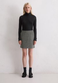 Schwarzer gerippter Rollkragenpullover, grauer Filz-Minirock, schwarze Schuhe und dunkle Socken. Schlichte, figurbetonte Silhouette mit glatter Textur.