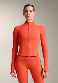Červená športová bunda s vysokým golierom a plným zipsom. Dlhé rukávy majú otvory pre palce. Navrhnutá s priliehavým strihom a hladkou textúrou.
