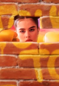 Une jeune femme portant des gants de boxe jaunes jette un coup d'œil à travers un trou dans un mur en briques recouvert de graffiti, avec une expression concentrée.