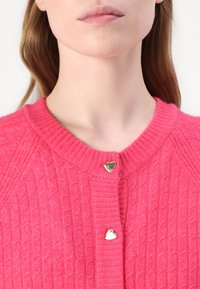 Rosa stickad cardigan med texturerat mönster, utrustad med tre guldhjärtformade knappar vid kragen för säker stängning.