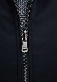 Svart zip-jacka med detaljer som inkluderar en metallisk dragkedja och ett mönstrat innerfoder med cirklar i blå och gräddtoner.