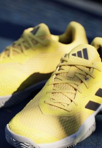 Zapatillas deportivas de color amarillo brillante con un upper texturizado, detalles en negro y un diseño aerodinámico. Suela de goma con un dibujo que proporciona agarre.