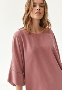 Vaaleanpunainen paita, jossa on väljä istuvuus, venepääntie ja kolme neljännespituista hihaa, joissa on röyhelöreunat. Sujuva kankaan rakenne.