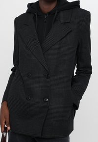 Blazer doppiopetto nero con un motivo houndstooth testurizzato, dotato di ampi rever e bottoni neri, indossato sopra una felpa con cappuccio.