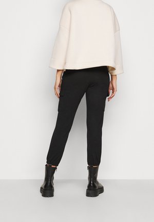 Personne debout, tournée de dos, portant un pantalon cargo noir, un pull oversize couleur crème et des bottines noires épaisses sur un fond uni.