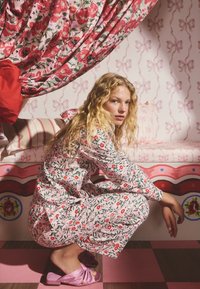Floral-print pink pajama set features a mix of red and green patterns. The model sits on a pink checkered floor, wearing pink bow-accented slides.