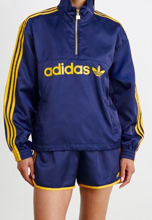 Osoba nosząca granatowy dres Adidas z żółtymi paskami, składający się z kurtki z półzamkiem i dopasowanych szortów z żółtą lamówką.