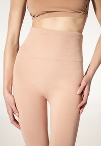 Kvinna klädd i högmidjade, ribbad, rosa leggings och matchande croppad topp, stående mot en ljus bakgrund.