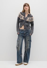 Cardigan grigio lavorato a maglia con motivi floreali crema e grigio scuro, abbinato a jeans cargo blu con multiple tasche e vestibilità comoda.