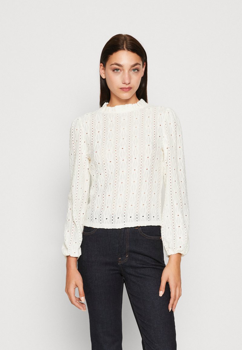 Balta garās piedurknes blūze ar teksturētu audumu, ar ruffled kakla izgriezumu un caurumiņu detaļām, savienota ar tumšām džinsu biksēm.