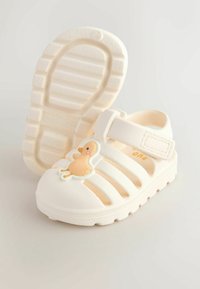 White rubber sandals with ventilated design, featuring a duckling applique on the front and a velcro strap for secure fit. Textured sole.