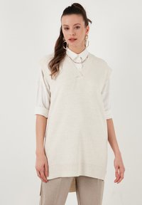 V-hals ærmeløs strikvest i lys beige med struktureret finish og ribbet kant, lagdelt over en hvid knap-op skjorte.