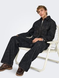 Donker denim jumpsuit met contrasterende gele stiknaden, ritssluiting en twee borstzakken; gedragen met bruine veterlaarzen en zittend op een stoel.