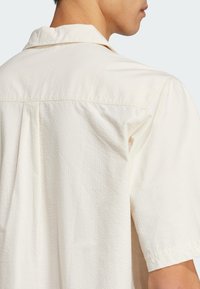 Short-sleeve, light cream shirt featuring a textured pattern and a camp collar, with a visible back pleat for added detail.