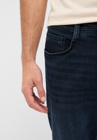 Ciemne jeansy z denimu o subtelnej teksturze gradientowej; posiadają boczną kieszeń, a obok niej spoczywa ręka.