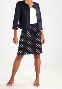 Veste en daim marine à manches trois quarts, portée sur un haut blanc. Jupe noire avec des pois blancs et un ourlet festonné. Sandales marron clair.