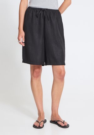 Person iført sorte løse knælange shorts med snøre i taljen, grå ærmeløs top og sorte flip-flops står på hvidt gulv.