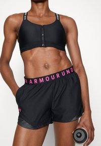 Idrottsperson i svart sport-BH med dragkedja fram och svarta shorts med rosa Under Armour-logga, håller en vattenflaska i höger hand.