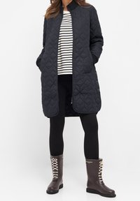 Pletený tmavě modrý dlouhý kabát s černým žebrovaným límcem, nošený přes pruhované tričko, černé legíny a hnědé voděodolné šněrovací boty.